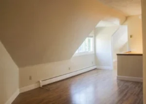 An empty room with a sloping ceiling, wooden floor, and a window. The room is well lit.